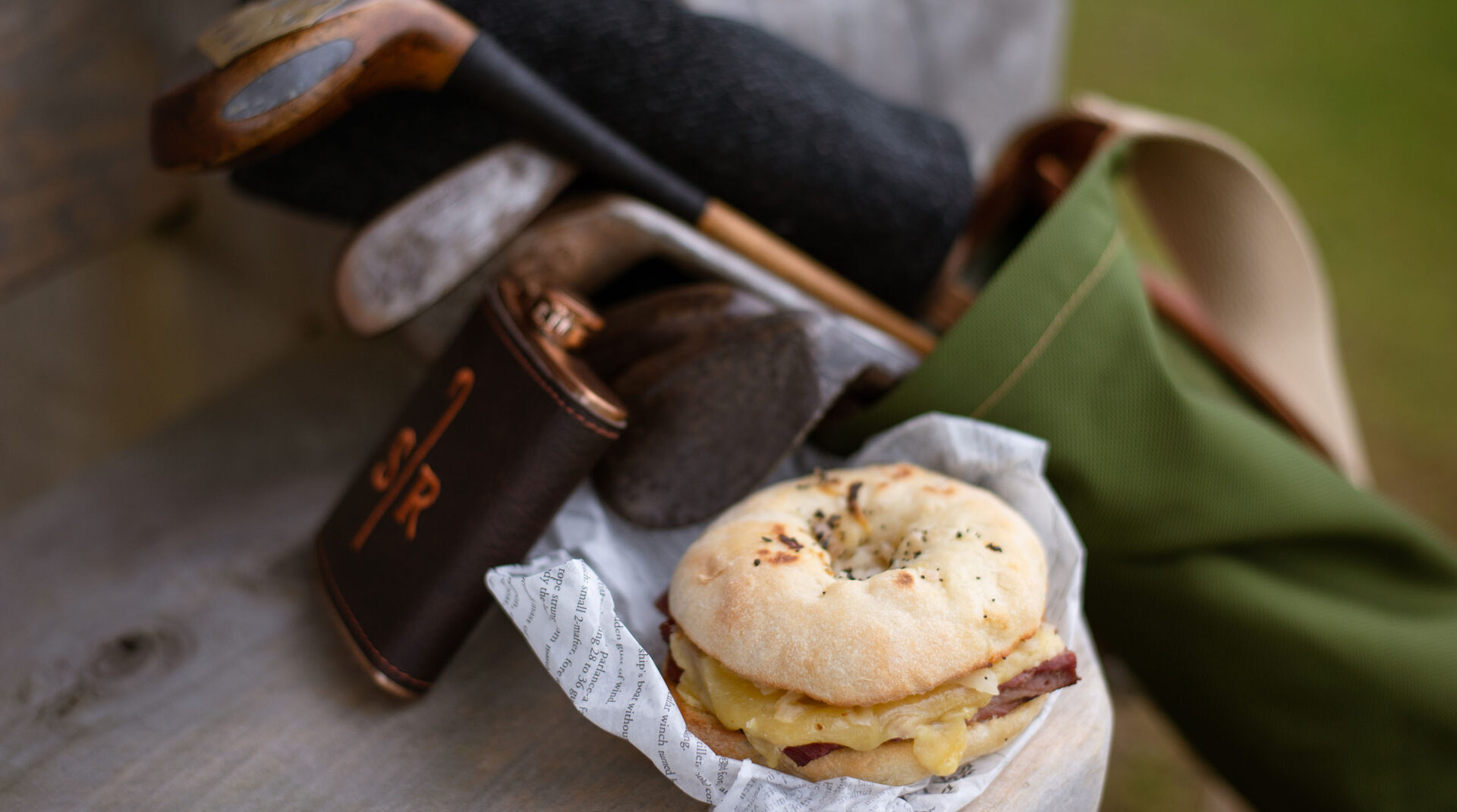 Breakfast bagel nestled on butcher paper next to a leather wrapped flask and golf clubs